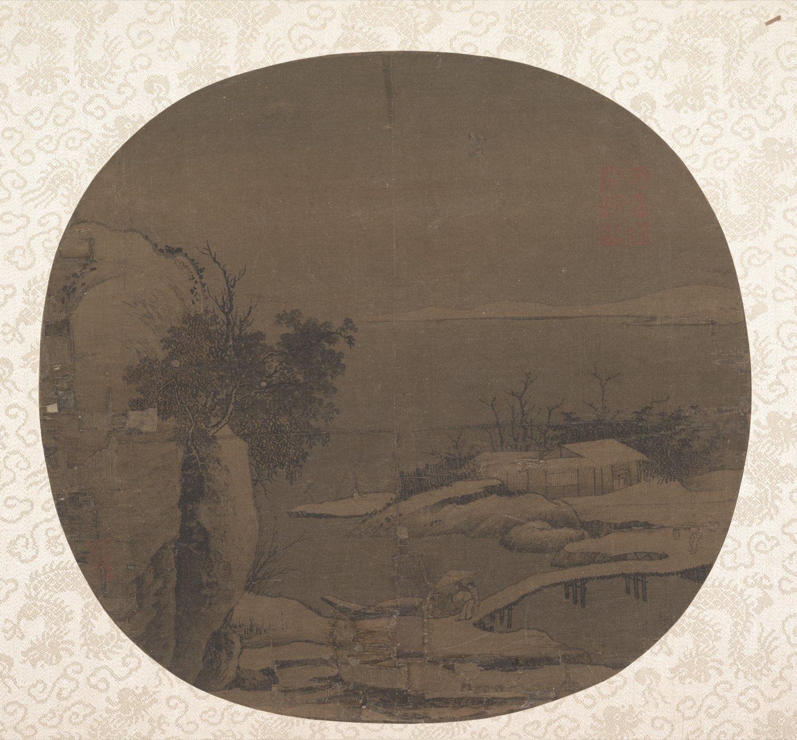 一幅深色绢本团扇,衬在浅色花纹的裱绫上。左边是一段长着斜树的岩岸;雪落在岸边低矮的屋顶、石头和一座小木桥上,宽阔平静的河面一直延伸到对岸隐约的山。